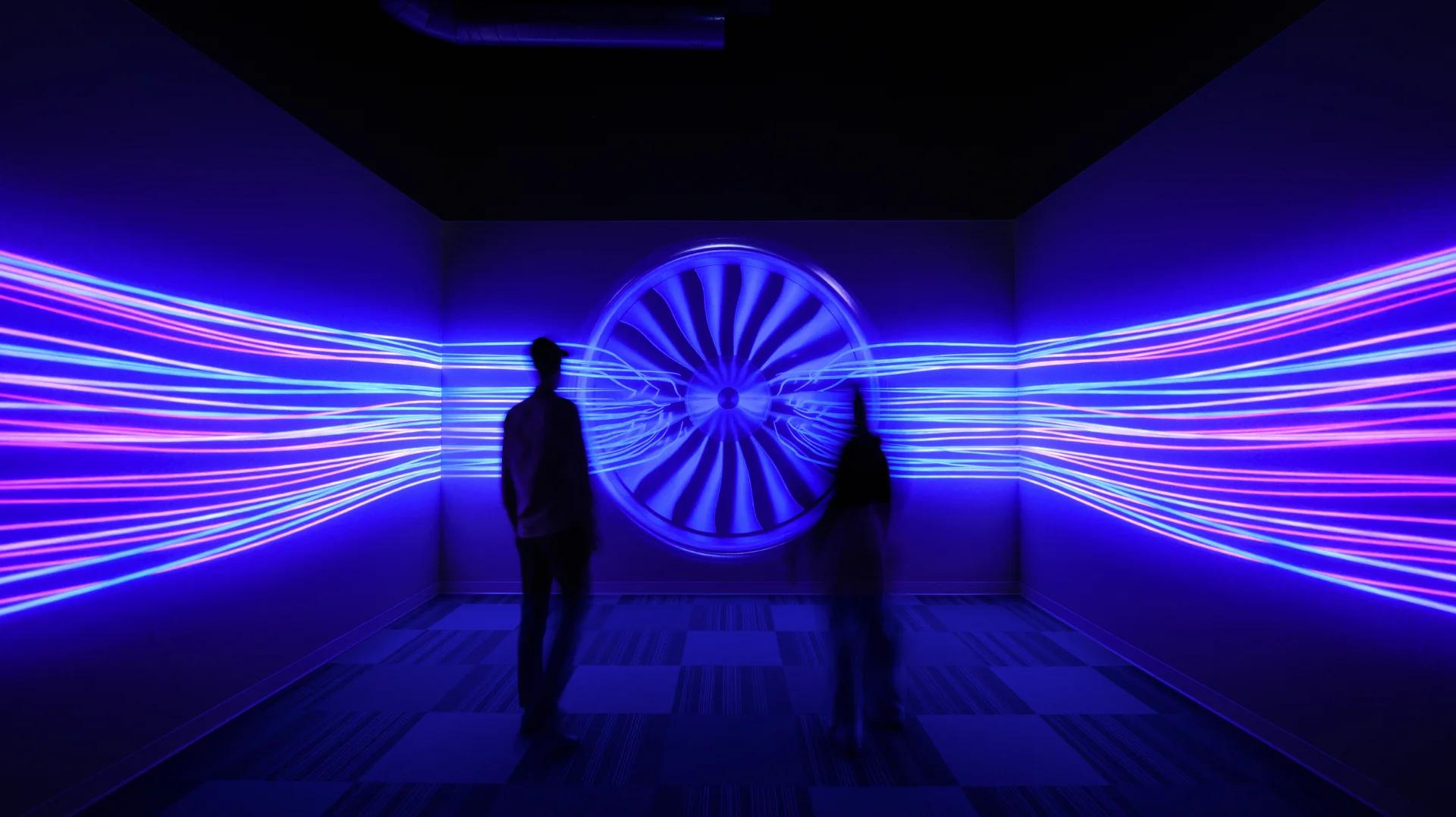 two people standing in the center of a small room with a glowing blue image of an airplane engine covering all three walls